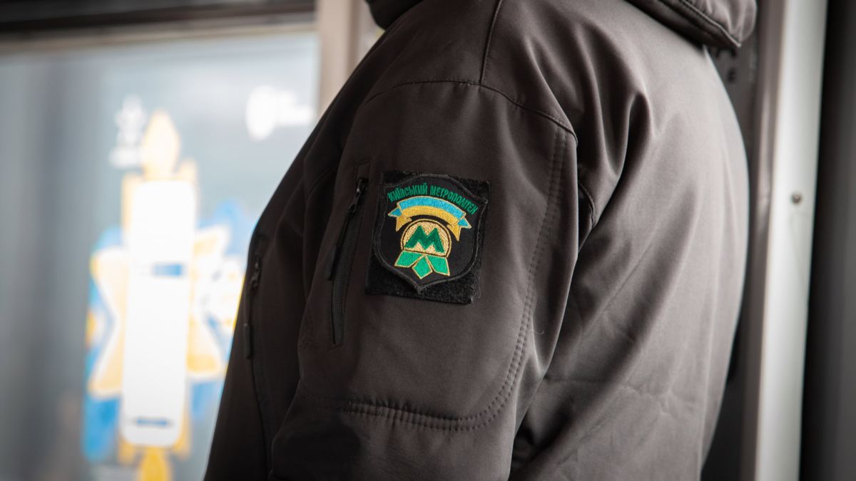 Працівник Київського метрополітену у темній куртці з нашивкою-шевроном підприємства на рукаві, на фоні вагона метро