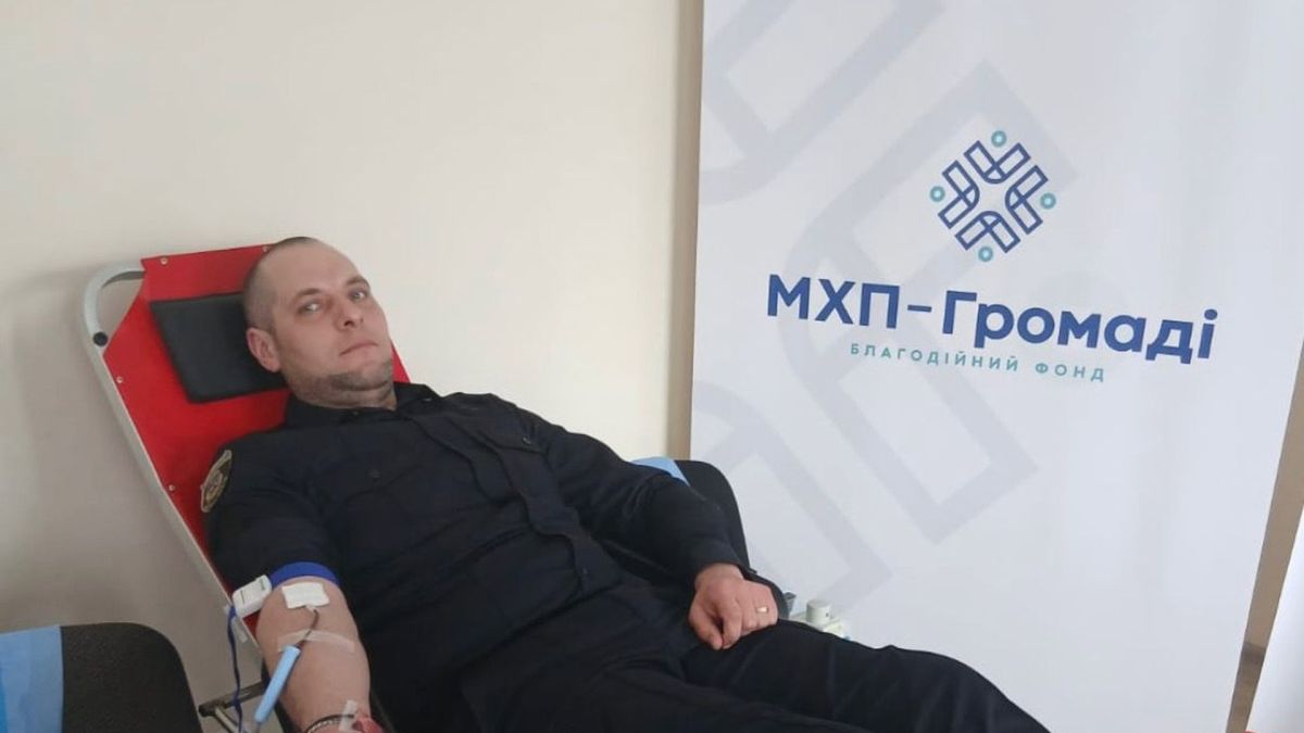 Поліцейський у формі здає кров під час донорської акції на підтримку поранених військових, банер благодійного фонду МХП-Громаді на фоні
