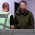 Головні новини Києва | Kyiv.News Поліцейський стоїть поруч із затриманим чоловіком у приміщенні відділку під час розслідування справи про вимагання 15 000 доларів за вплив на ВЛК