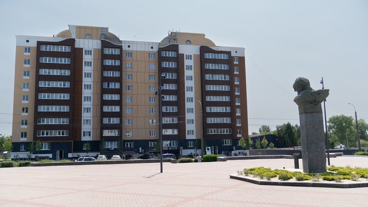 Сучасна багатоповерхівка з коричневими й бежевими фасадами, перед якою на площі встановлений пам’ятник у вигляді бюста на високому постаменті
