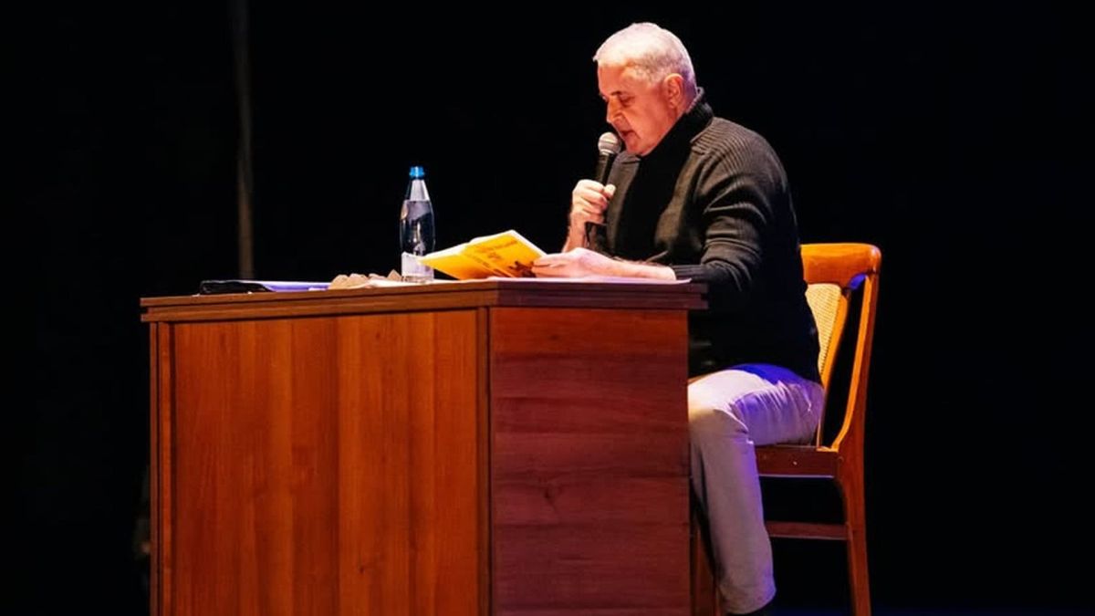 Лесь Подеревʼянський сидить на стільці біля деревʼяного стола, він із мікрофоном читає текст свого виступу в темній залі