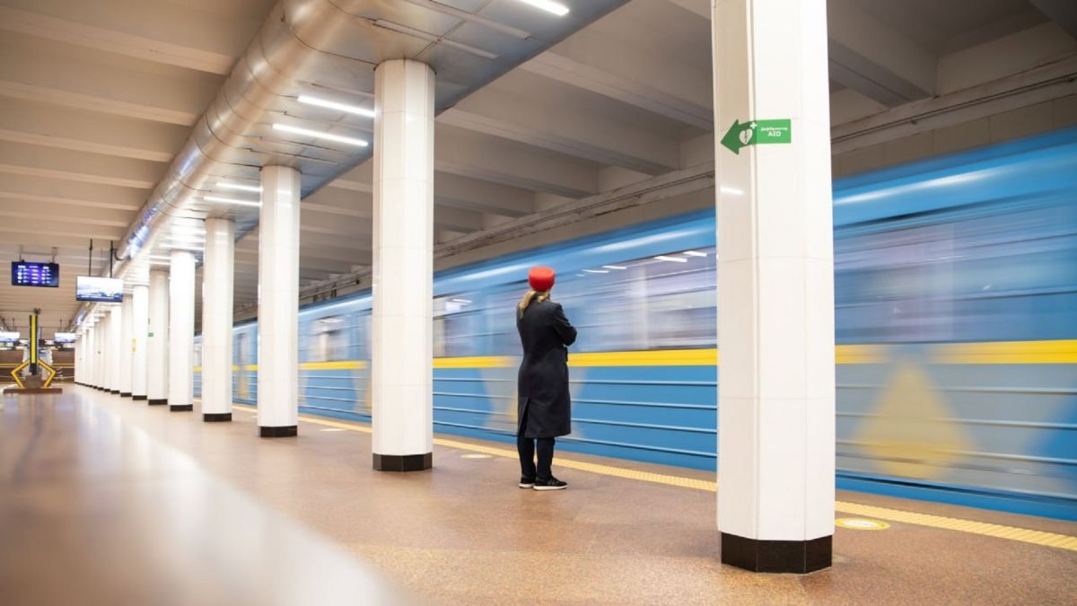 Синьо-жовтий потяг Київського метрополітену проїжджає повз станцію, пасажирка чекає на платформі