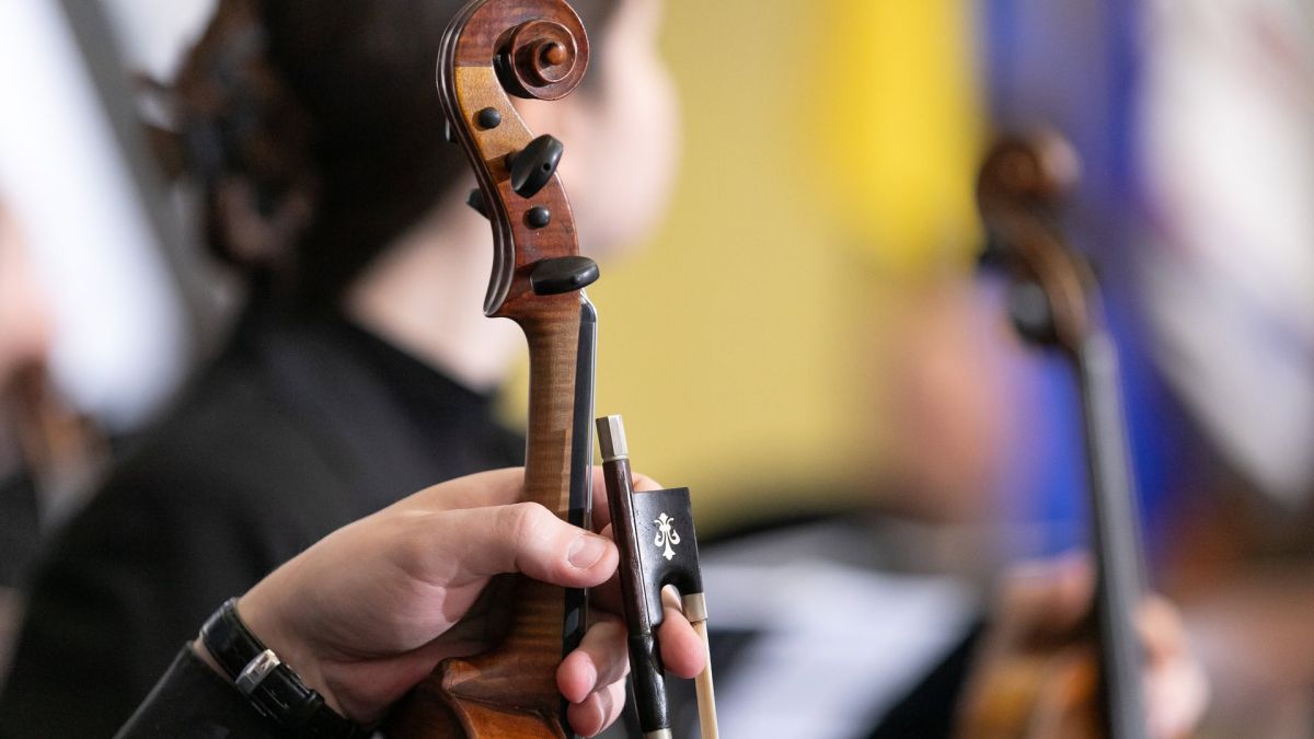 Рука музиканта тримає скрипку та смичок крупним планом на тлі українського прапора під час концерту в Києві