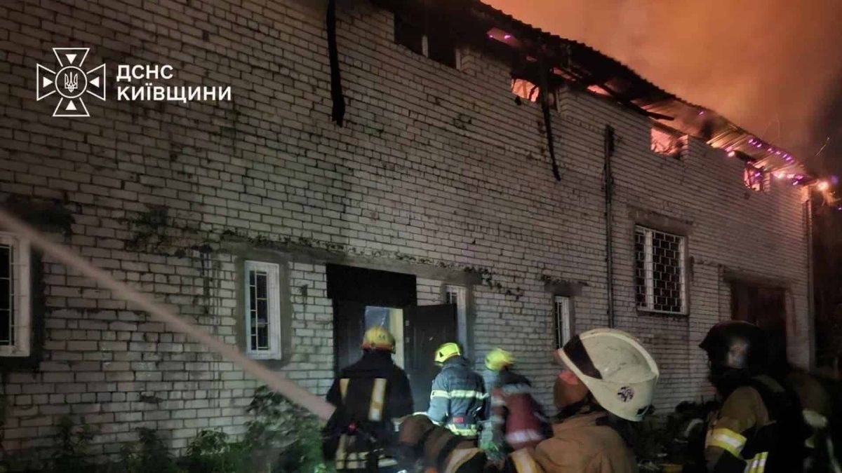 Група рятувальників ДСНС Київщини стоїть біля входу до цегляної будівлі, дах якої охоплений полум'ям