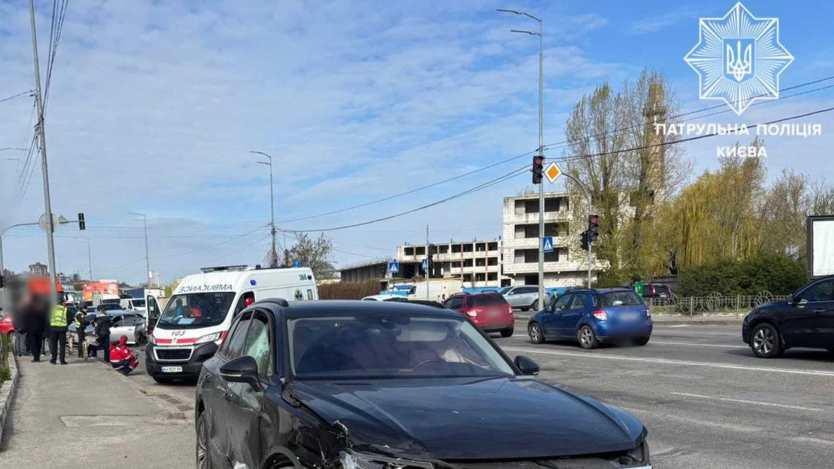 Пошкоджений чорний позашляховик після ДТП на перехресті Набережно-Лугової та Оленівської вулиць у Києві — зім'ятий передній бампер, поруч карета швидкої допомоги